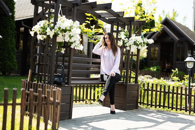 De jonge mooie donkerbruine vrouw in overhemd zit op houten bank met bloemen in tuin