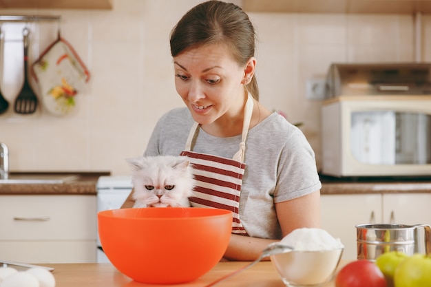 De jonge mooie gelukkige vrouw met een witte Perzische kat bereidt deeg voor taarten met tablet op tafel in de keuken. Thuis koken. Eten koken.