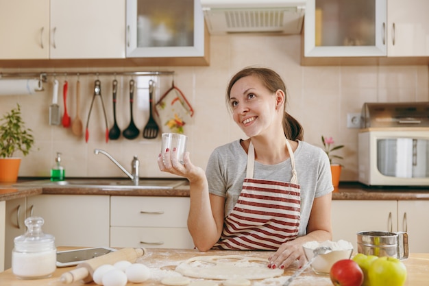 De jonge mooie gelukkige vrouw zit aan een tafel met bloem en knipt een glas uit in deegcirkels voor knoedels in de keuken. Thuis koken. Eten koken.