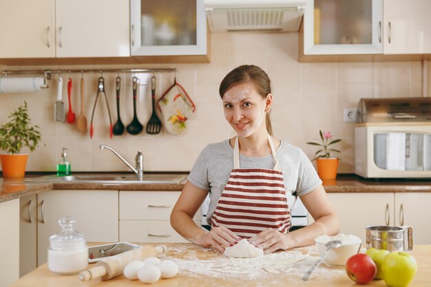 De jonge mooie gelukkige vrouw zit aan een tafel met bloem, kneed deeg en gaat een taart in de keuken bereiden. Thuis koken. Eten koken.