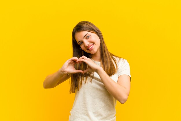 De jonge mooie vrouw die en gelukkig, leuk, romantisch en in liefde glimlachen voelen, maakt hartvorm met allebei overhandigt oranje muur