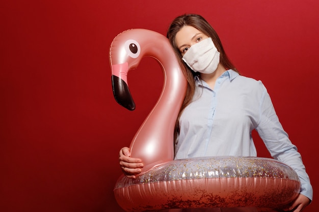 De jonge mooie vrouw van de bureauwerknemer op rode achtergrond in beschermend medisch masker koestert zwemmende cirkel roze flamingo in pandemie, vakantieannulering