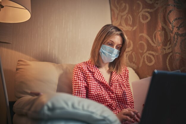 De jonge onderneemster in een medisch beschermend masker werkt vanuit huis bij de computer tijdens zelfisolatie en quarantaine.