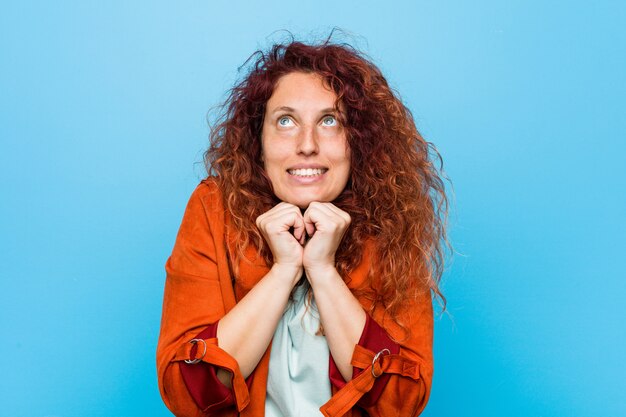 De jonge roodharige elegante vrouw houdt handen onder kin, kijkt gelukkig opzij.