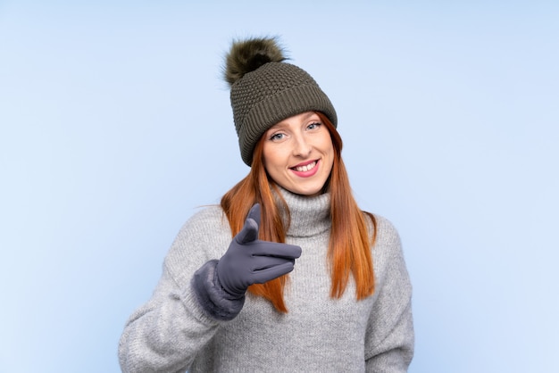 De jonge roodharige Russische vrouw met de winterhoed over geïsoleerde blauwe achtergrond richt vinger op u