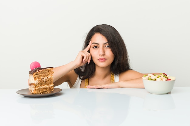 De jonge Spaanse vrouw die tussen cake of fruit kiezen die zijn tempel met vinger richten, het denken, concentreerde een taak.