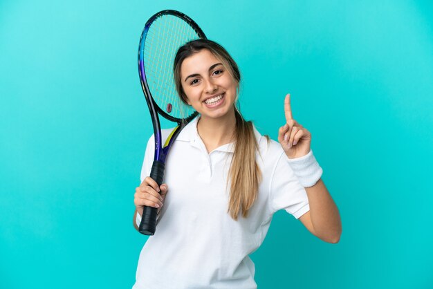 De jonge speler van het vrouwentennis die op blauwe muur wordt geïsoleerd die en een vinger in teken van het beste toont opheft