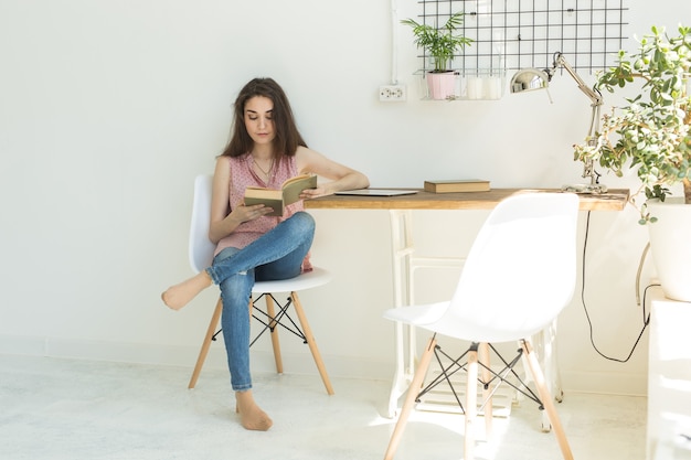 De jonge studentenvrouw leest een boek, terwijl zij op witte stoel in een witte ruimte zit