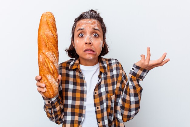 De jonge vrouw die brood maakt dat op witte muur wordt geïsoleerd, haalt schouders op en opent verwarde ogen