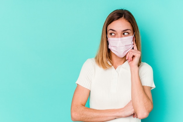 De jonge vrouw die een masker voor virus draagt dat op blauwe muur wordt geïsoleerd, ontspant het denken over iets dat een exemplaarruimte bekijkt.