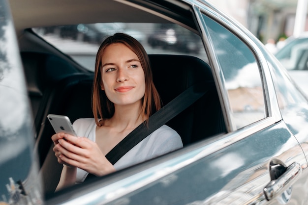 De jonge vrouw die een smartphonezitting in de auto houden kijkt uit in het venster