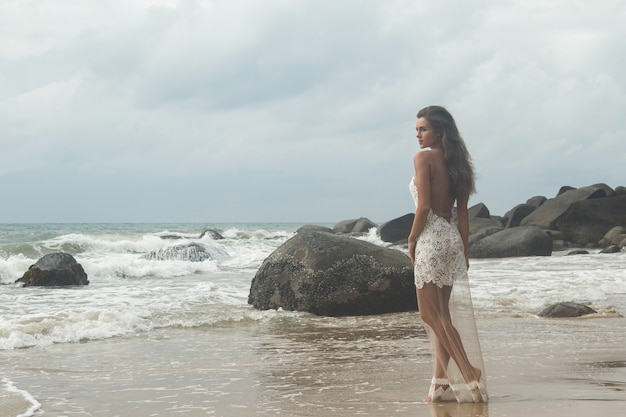 De jonge vrouw die mooie witte kleding draagt, stelt op het rotsachtige strand