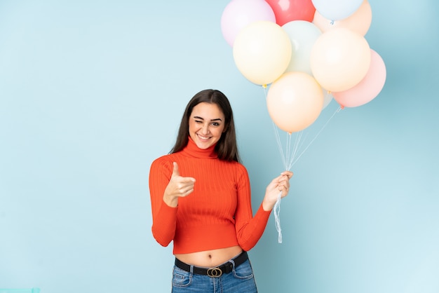 De jonge vrouw die vele ballons vangt die op blauwe muur worden geïsoleerd wijst vinger op u