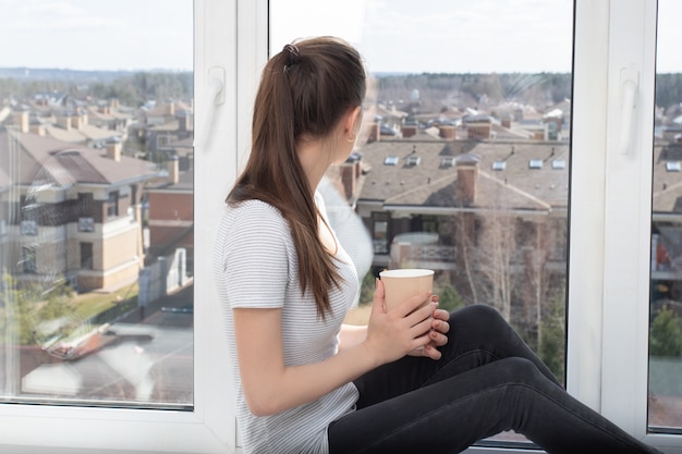 De jonge vrouw drinkt een hete drank terwijl thuis het zitten op een venster