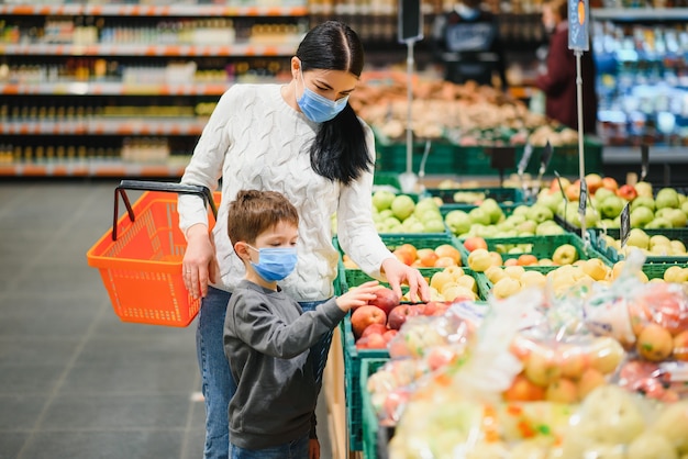 De jonge vrouw en haar zoontje die beschermend gezichtsmasker dragen winkelen een levensmiddel in een supermarkt