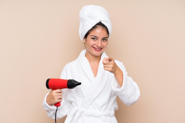 De jonge vrouw in een badjas met haardroger over geïsoleerde muur richt vinger op u met een zekere uitdrukking