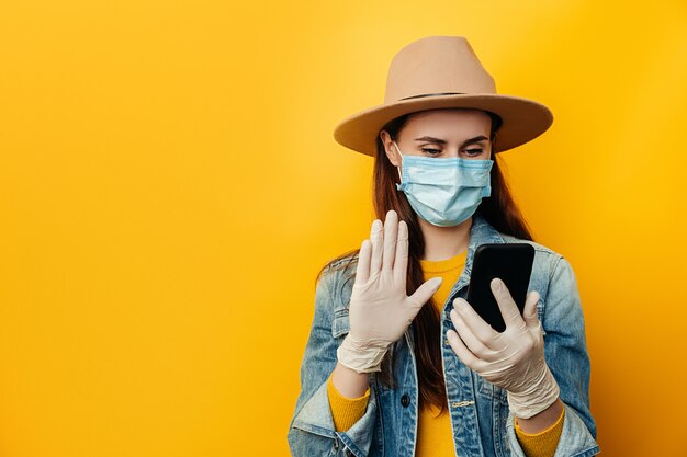 De jonge vrouw in medische steriele handschoenen van het gezichtsmasker, die smartphone gebruiken, met vrienden babbelen, e-mail met goed nieuws lezen, surfen op het web, draagt denimjasje en hoed, die over gele achtergrond wordt geïsoleerd.