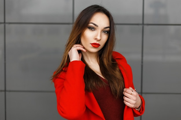 De jonge vrouw in rode kleren stelt dichtbij de grijze muur