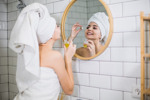 De jonge vrouw in witte handdoek past vochtinbrengende olie op gezichtshuid toe. Zelfzorg, schoonheid en huidverzorging concept.