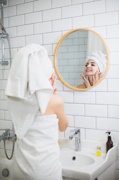 De jonge vrouw in witte handdoek past vochtinbrengende olie op gezichtshuid toe. Zelfzorg, schoonheid en huidverzorging concept.