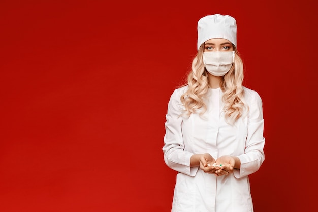 De jonge vrouw in witte medische laag en medisch masker houdt handvol pillen, die bij de rode achtergrond met exemplaarruimte worden geïsoleerd. Concept van gezondheidszorg en ziektepreventie.