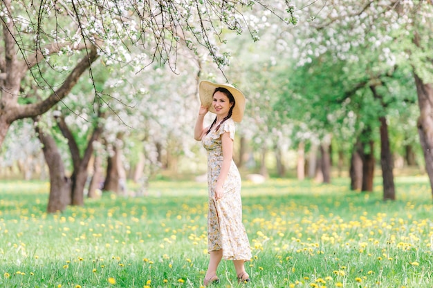 De jonge vrouw met strohoed loopt in de gebloeide tuin. Meisje onder Sakura-bloesemboom. Zomer mode concept