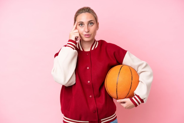 De jonge vrouw van de basketbal kaukasische speler die op roze achtergrond wordt geïsoleerd die een idee denkt