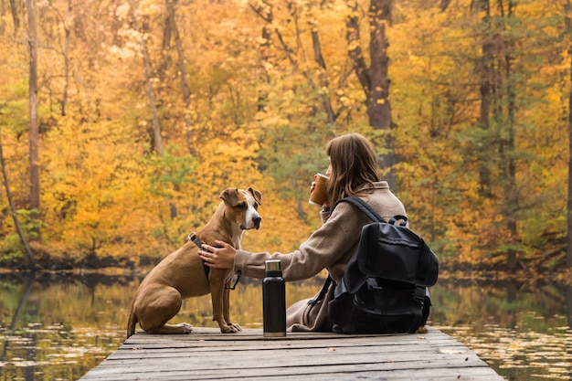 De jonge vrouw van de toeristenwandelaar zit bij rivieroever met haar huisdierenpuppy en geniet van mooi nazomerzicht met kop van koffie van thermosflessen