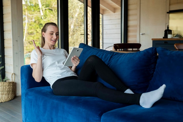 De jonge vrouw zit op een blauwe bank in een nieuw huis met behulp van een tablet en viert een prestatie w