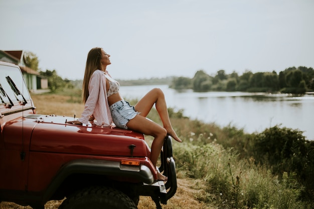 De jonge vrouw zit op een rode autokap en geniet van in de zomerzon