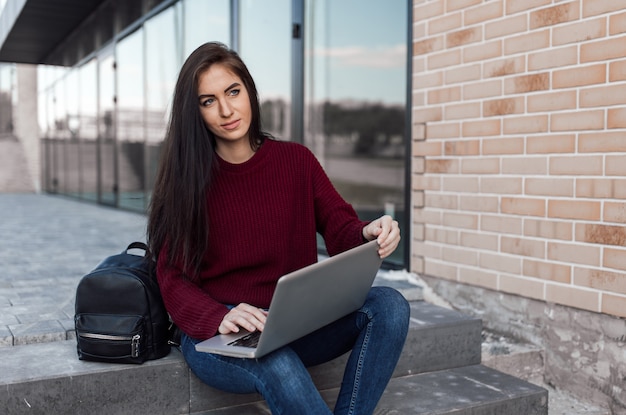 De jonge vrouw zit op stedelijke treden en werkt met laptop op straat