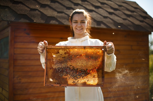 De jonge vrouwelijke imker houdt houten frame met honingraat