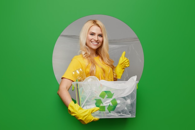 De jonge vrouwelijke vrijwilliger in gele handschoenen met recyclingsbak met plastic afval kijkt door het ronde gat op een groene achtergrond. Bescherming van het milieu concept