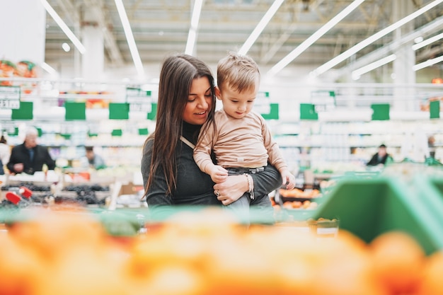 De jonge vrouwenmoeder met leuk de peuterkind van de babyjongen op handen koopt de verse vruchten in supermarkt