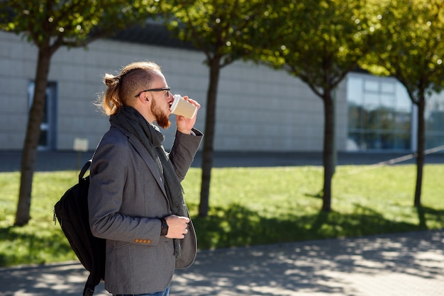 De jonge zakenman met rugzak drinkt koffie in openlucht tijdens lunchpauze dichtbij modern bureaucentrum. Beambte op een werkende lunchpauze.
