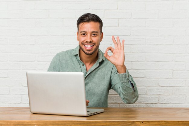 De jonge zitting die van de filipijnse mens met zijn laptop werkt vrolijk en zeker tonend ok gebaar.