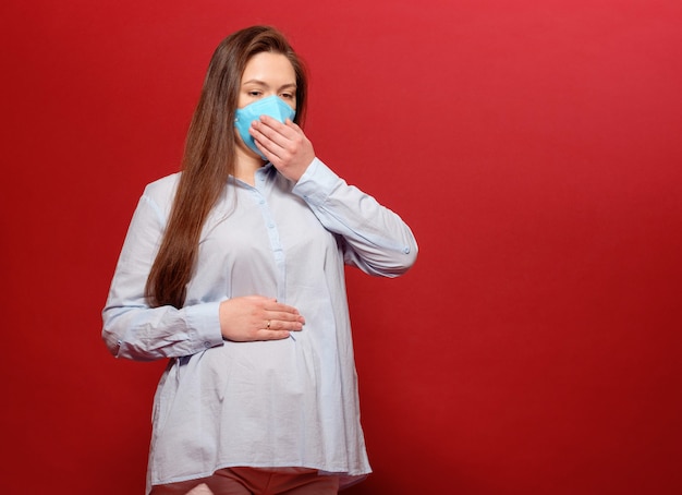 De jonge zwangere vrouw op rode muur in beschermend medisch masker houdt op maag en niest