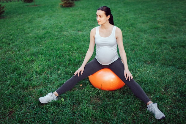 De jonge zwangere vrouw zit op oranje fintess bal met uitgespreide benen in park. Ze kijkt naar rechts.