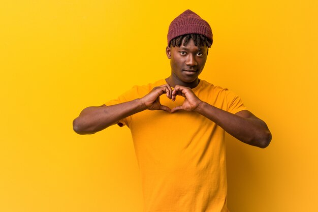 De jonge zwarte mens die rastas over geel draagt glimlacht en toont een hartvorm met hem handen.