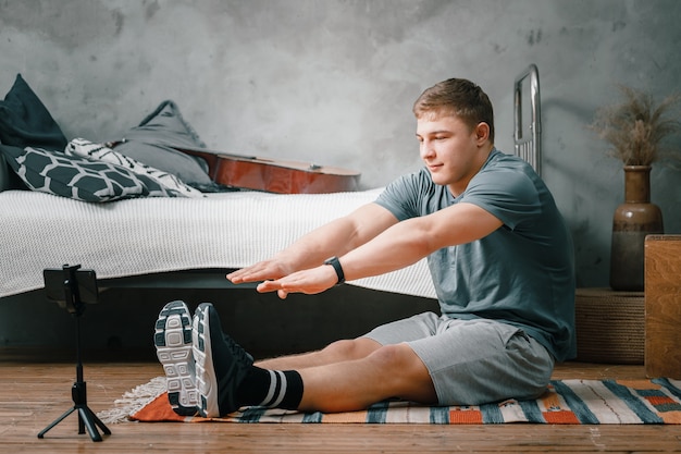 De jongeman gaat thuis sporten. Sportman met blond haar strekt zich uit met vingers in sokken op tapijt in de slaapkamer