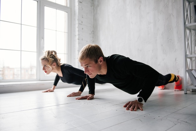 De jongen en het meisje in trainingspak. Zwart sportuniform. Mannelijke en vrouwelijke atleten. Opgepompt lichaam. Opdrukken. Ochtendtraining. Set oefeningen voor lichaam. Klassen in paren. Samen thuis trainen.