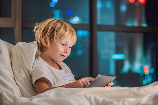 De jongen gebruikt de tablet in zijn bed voordat hij gaat slapen op een achtergrond van een nachtstad. Kinderen en technologieconcepten