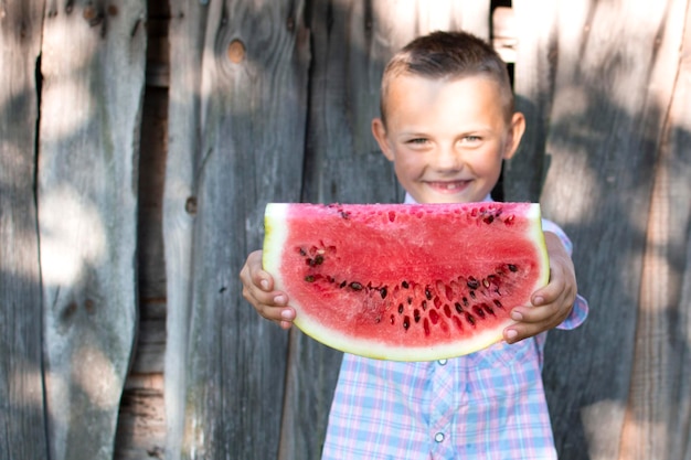 De jongen houdt een groot stuk van watermeloen tegen een houten landelijke muur