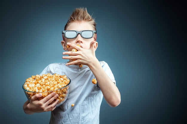 De jongen houdt in zijn handen en eet popcorn kijken naar een film in 3d-bril, blauwe muur. het concept van een bioscoop, films, emoties, verrassing, vrije tijd. streaming platforms.