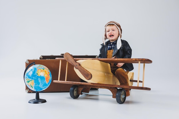 Foto de jongen huilt in een leren jas en een pilotenmuts een houten vliegtuig bruine koffers houten speelgoed voor kinderen baby emoties