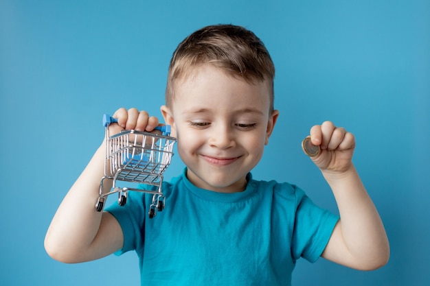 De jongen in blauwe t-shirt houdt boodschappenwagentje en muntstuk op blauw.