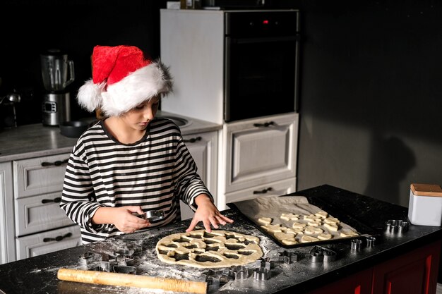 De jongen in de hoed van de Kerstman bakt een Kerstmiscake in de keuken. In zwart, rood en wit tinten