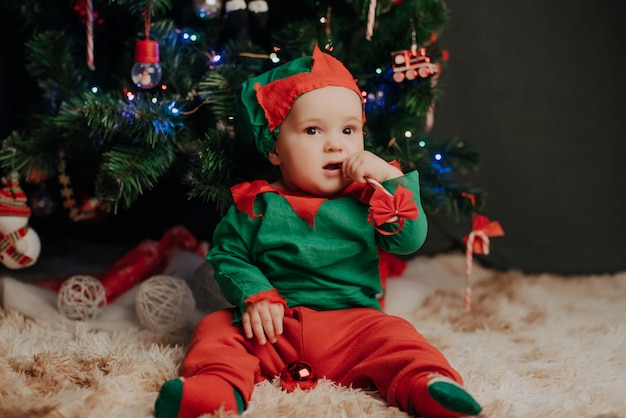 De jongen in een elfkostuum zit onder een kerstboom met een lolly