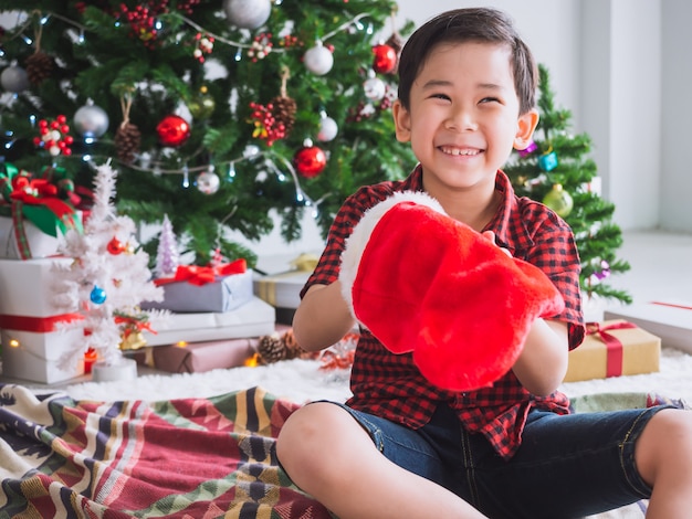 Foto de jongen in een rood overhemd houdt rode sok en gelukkig met grappig om kerstmis met kerstmisboom te vieren