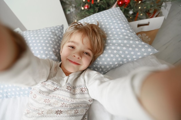 De jongen in pyjama's voorschoolse leeftijd zit op bed en fotografeert zichzelf.